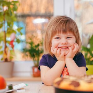 A happy young girl smiling indoors surrounded by plants and a pastry, with natural light streaming in.